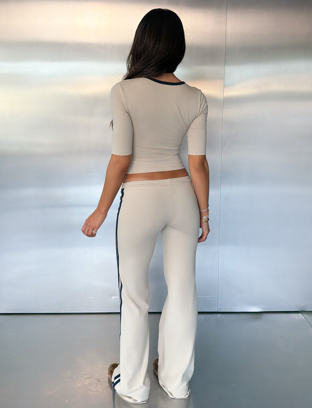 A woman stands indoors facing a metallic wall, wearing the SHANICE TOP in sand by TIGERMIST—a bodycon three-quarter sleeve top—paired with matching flare pants featuring dark side stripes.