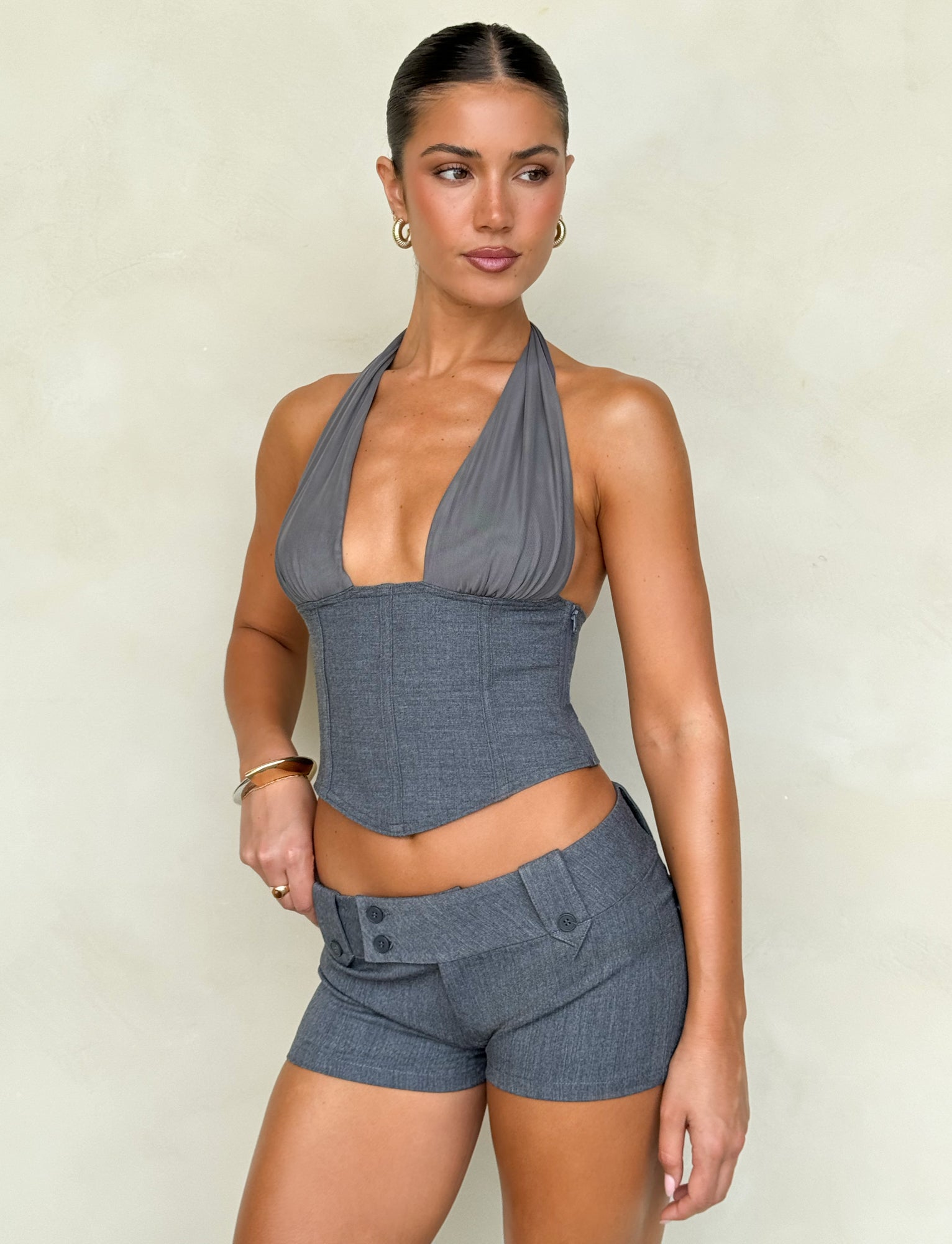 A person stands confidently against a light background, dressed in the chic JANECE TOP - GREY from TIGERMIST. They wear a gray halter top with a plunging neckline, paired with matching shorts. Their hair is styled in an elegant updo and accented by hoop earrings, while one hand rests assertively on their hip.