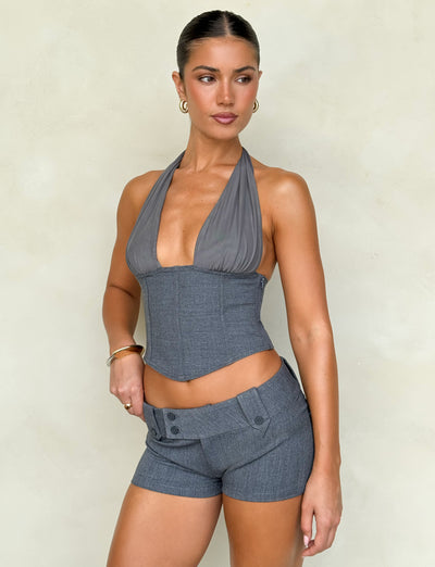 A person stands confidently against a light background, dressed in the chic JANECE TOP - GREY from TIGERMIST. They wear a gray halter top with a plunging neckline, paired with matching shorts. Their hair is styled in an elegant updo and accented by hoop earrings, while one hand rests assertively on their hip.