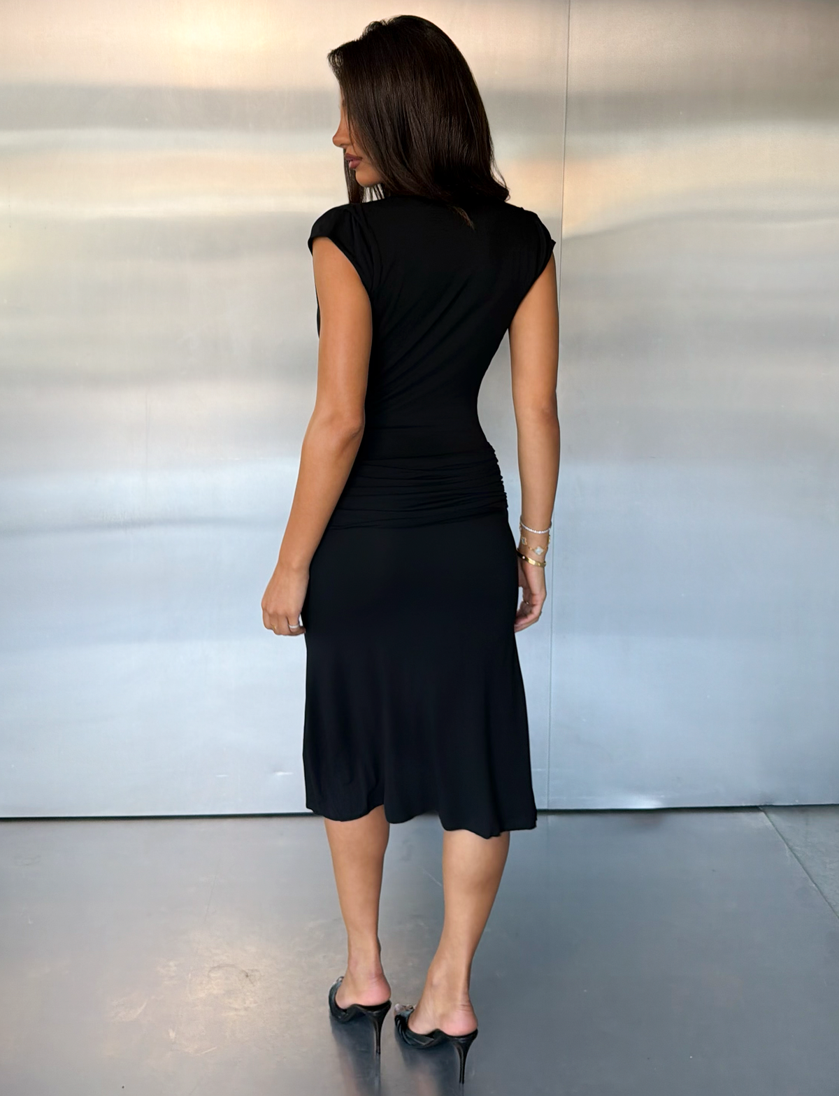 A woman with long brown hair, wearing the TIGERMIST ARIELLA PLUNGE MIDI DRESS in black and black heels, stands indoors facing away from the camera in front of a gray wall.