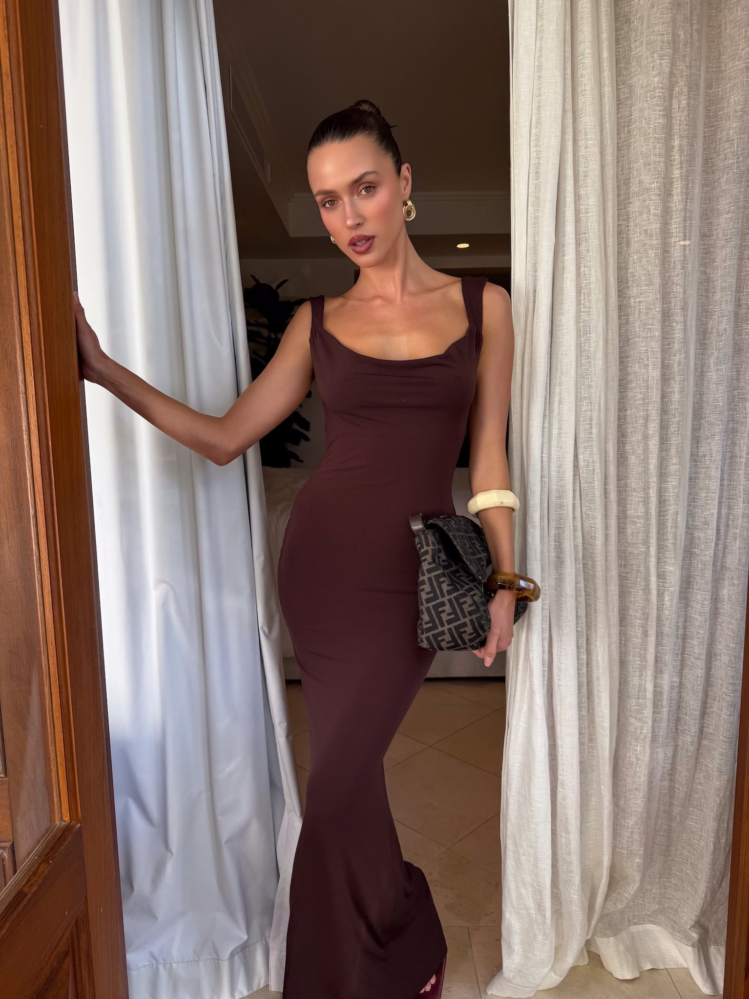 A woman stands in a doorway, wearing the TIGERMIST MORANA DRESS - BROWN : CHOCOLATE, accessorized with a patterned clutch and bold bracelets, her hair in a sleek updo between flowing white curtains.