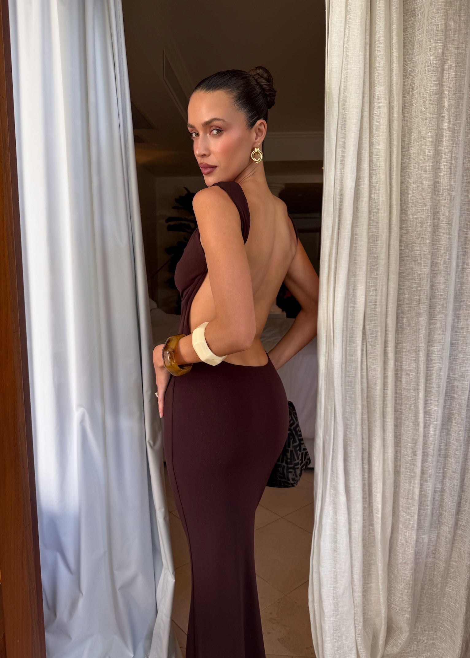 A woman with dark hair in a bun poses between white curtains, wearing the TIGERMIST MORANA DRESS - BROWN : CHOCOLATE, a bodycon maxi dress, with gold earrings. She looks over her shoulder toward the camera in a bright room.