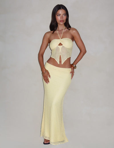 A woman with long dark hair is posing against a plain background, confidently showcasing the TIGERMIST SHAH SKIRT in yellow lemon. Her light yellow, form-fitting crochet two-piece outfit includes a halter top with intricate front details and a stylish maxi length skirt. She accentuates her look with bracelets and rings.