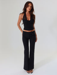 A woman with long dark hair stands against a plain background, wearing a black halter top paired with TIGERMIST's VESPER PANT in black. The fitted pants feature a low-rise waist. She completes her look with one arm behind her back, the other resting at her side, and black open-toed shoes.