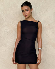 A woman models the TIGERMIST FREYA DRESS in black—featuring a buttery soft fabric and adjustable asymmetrical neckline—paired with silver hoop earrings and a chunky bracelet. Her dark hair is styled in a sleek bun as she gazes at the camera.