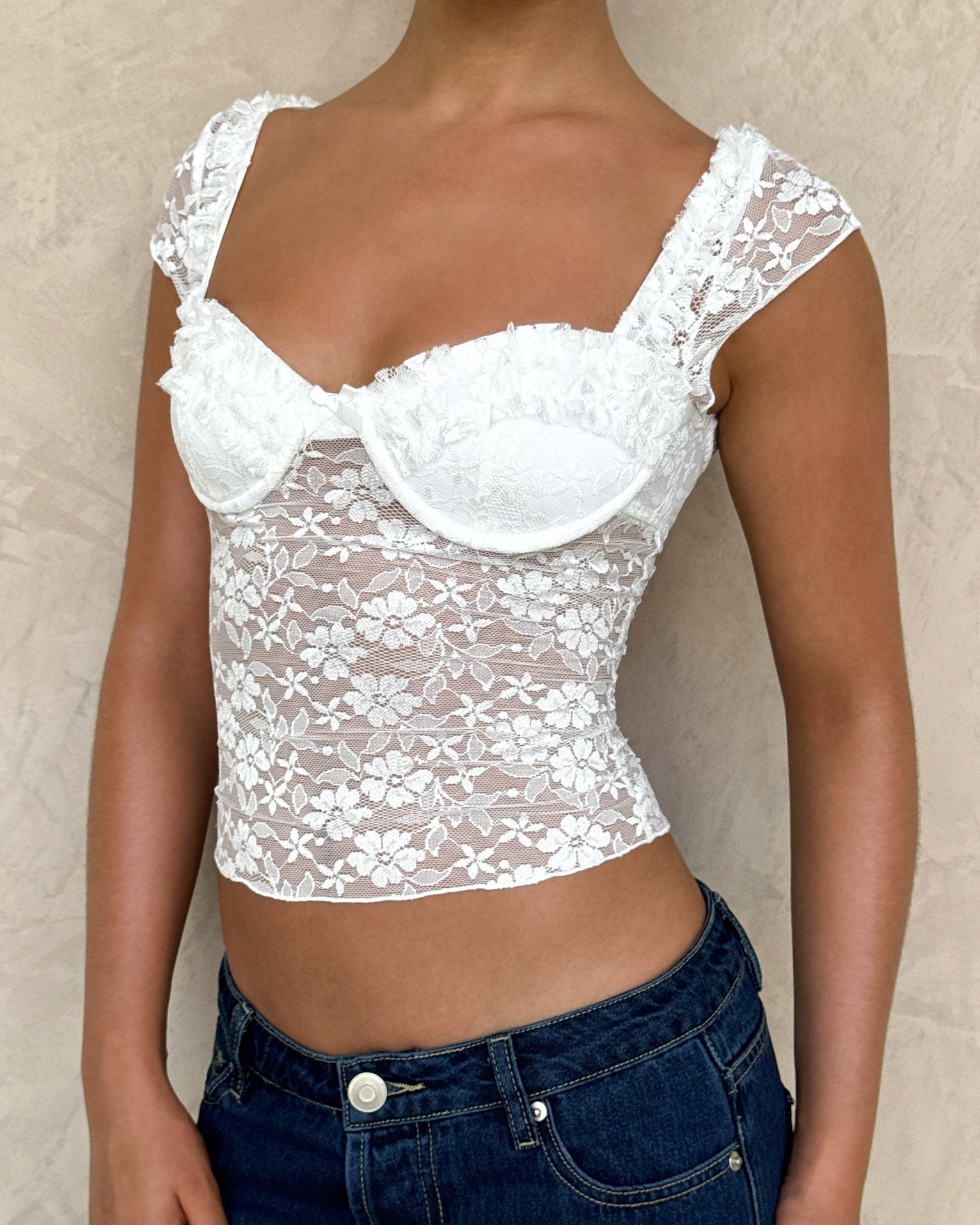 A person wears the NYRA TOP - WHITE by TIGERMIST, a white lace semi-sheer bustier with cap sleeves, floral patterns, and structured cups, paired with blue jeans against a beige textured wall.