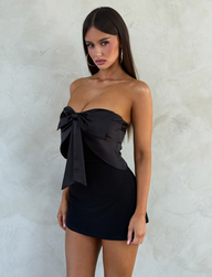 A woman with long dark hair wears the TIGERMIST GROVE MINI DRESS - BLACK, a strapless satin mini dress featuring a large bow on the chest. She stands against a light textured wall, gazing at the camera with a neutral expression.