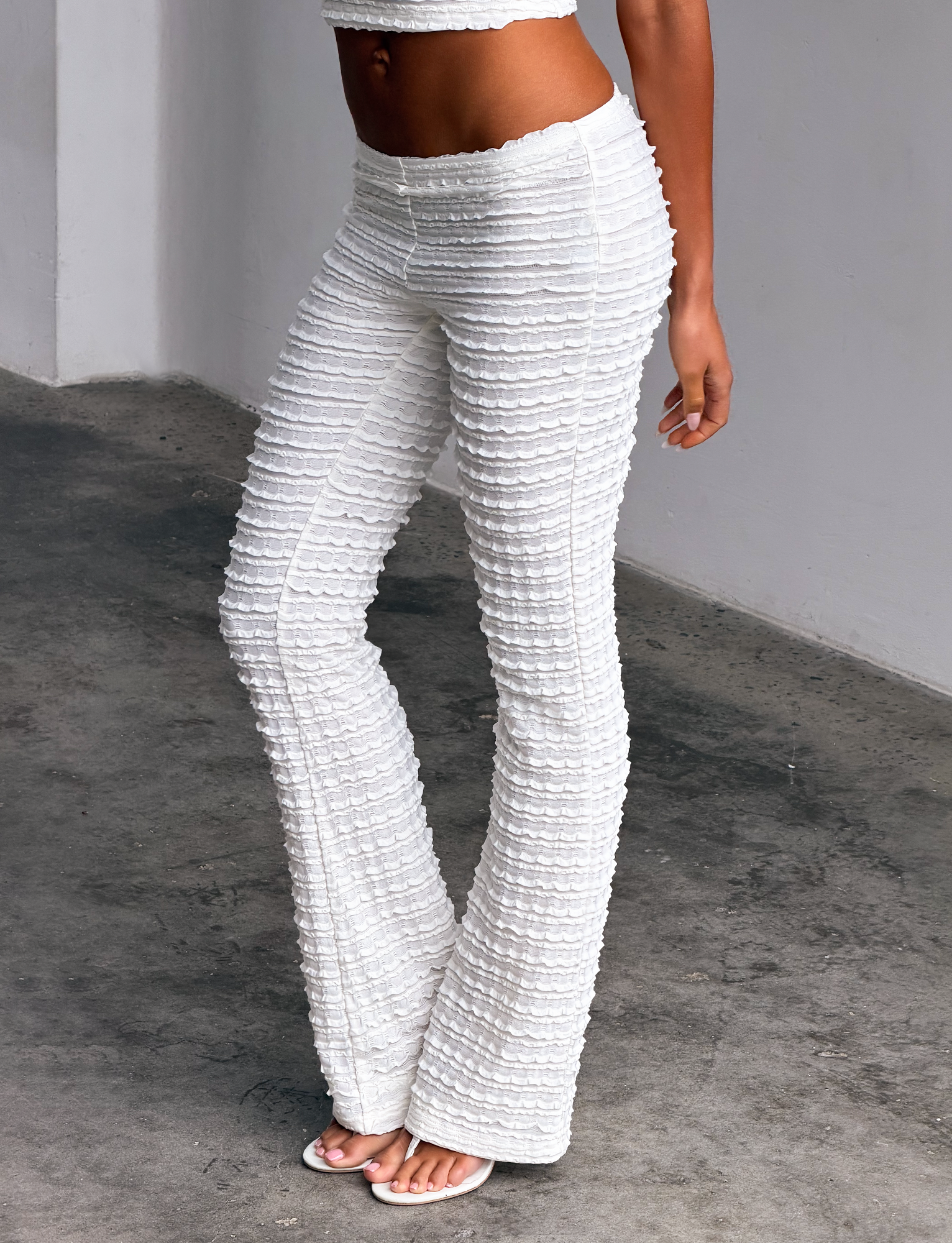 A woman stands indoors on a gray concrete floor, showing only her lower body while wearing TIGERMIST’s ZORA PANT in cream, styled with a matching crop top and white sandals against a light wall background.