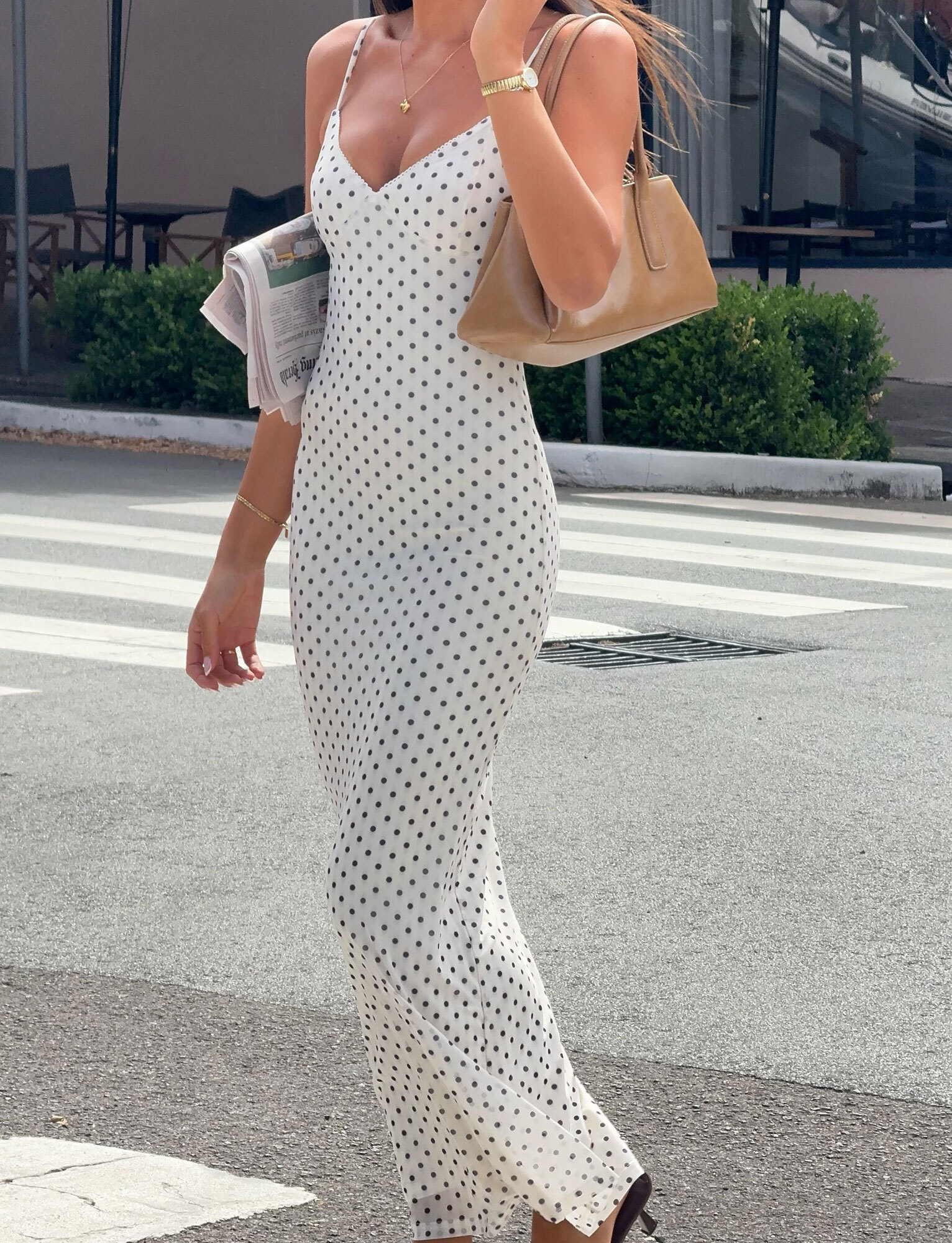 A woman wearing the TIGERMIST JULIANNA MAXI DRESS - POLKA DOT with adjustable straps walks on a city sidewalk, carrying a newspaper and tan handbag. She accessorizes with gold jewelry, brown sandals, and greenery with outdoor seating is in the background.