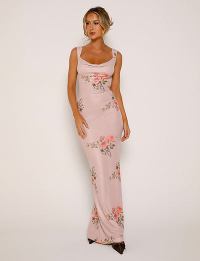 A person wearing the MORANA DRESS - PINK : FLORAL from TIGERMIST stands against a plain background. The long, form-fitting dress features a sleeveless design and a scoop neckline with elegant floral patterns. The individual has their hair styled in an updo and is looking slightly off to the side.