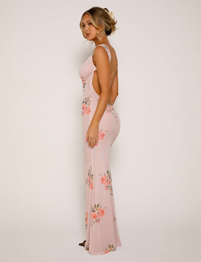 A woman stands against a plain background, wearing the MORANA DRESS - PINK : FLORAL from TIGERMIST. The long, form-fitting dress is adorned with floral patterns and features a low back and sleeveless design. She has blonde hair styled in an updo and is looking at the camera with a calm expression.