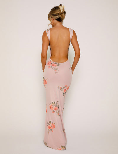 A woman is seen from the back wearing the MORANA DRESS - PINK: FLORAL by TIGERMIST, a long, light pink dress with a floral design. The dress features a deep open back and thick shoulder straps. Her hair is styled in a loose updo as she stands against a plain off-white background.