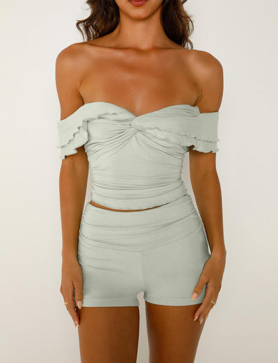 A woman is wearing the DENALI TOP - GREEN : SAGE GREEN from TIGERMIST with a ruffled, twisted top and matching high-waisted shorts. The background is plain white, and she has her hands resting at her sides. Her hair falls loosely over her shoulders.
