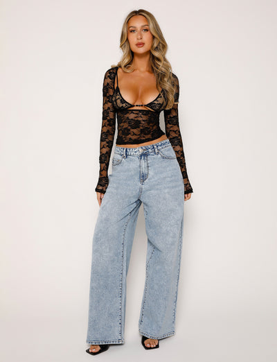 A woman stands against a plain background wearing the CHRISTY LONG SLEEVE TOP - BLACK : LACE from TIGERMIST and high-waisted, light-wash baggy jeans. She has long, wavy blonde hair and is accessorized with black open-toe heels. Her outfit seamlessly blends edgy and casual styles.