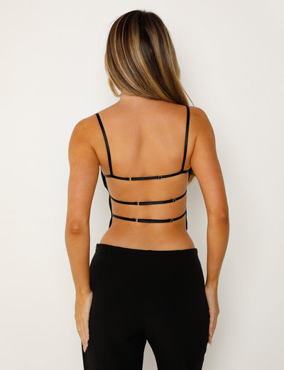 A person with long, straight hair is seen from behind, wearing the NAYSHA TOP - BLACK and black bottoms from TIGERMIST. The top has thin straps crossing horizontally across the back, leaving much of the back exposed. The background is plain white.
