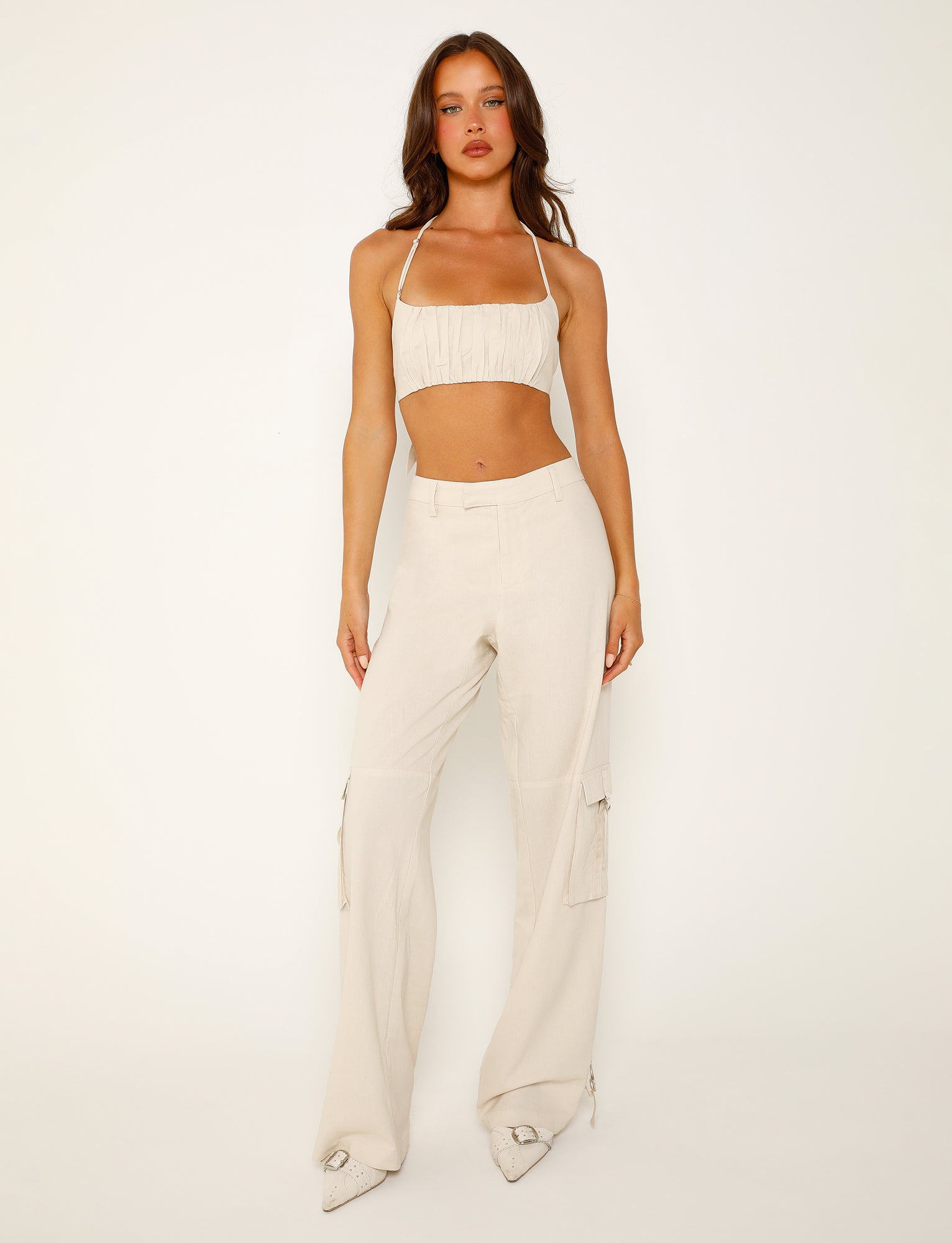 A person stands posing against a plain white background, showcasing a chic Tiger Mist ensemble. They are wearing a light beige crop top with thin straps, the KAYA PANT - WHITE : CREAM high-waisted cargo pants from TIGERMIST, and white pointed-toe shoes. The individual has long, wavy hair and a serious facial expression.