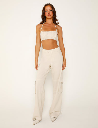 A person stands posing against a plain white background, showcasing a chic Tiger Mist ensemble. They are wearing a light beige crop top with thin straps, the KAYA PANT - WHITE : CREAM high-waisted cargo pants from TIGERMIST, and white pointed-toe shoes. The individual has long, wavy hair and a serious facial expression.