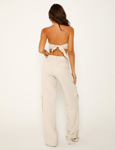 A woman with long, wavy hair poses with her back to the camera, showcasing a beige, backless halter top from Tigermist and matching high-waisted KAYA PANT - WHITE : CREAM cargo pants with side pockets. She completes the look with white high-heeled sandals against a plain white background.