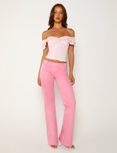A woman is posing against a plain white background, showcasing a stunning TIGERMIST ensemble. She is wearing an off-the-shoulder, ruffled pink top paired with the stylish KITTIE PANT - PINK. Completing her look are white high-heeled shoes. Her long hair is styled in loose waves, and she's looking confidently at the camera.