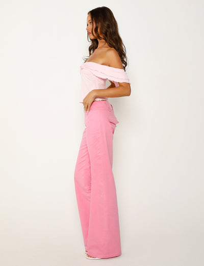 A woman stands in a side profile against a plain white background. She is wearing an off-the-shoulder light pink top and TIGERMIST's KITTIE PANT in pink. She has long, wavy hair and is looking forward. Her outfit is coordinated in shades of pink.