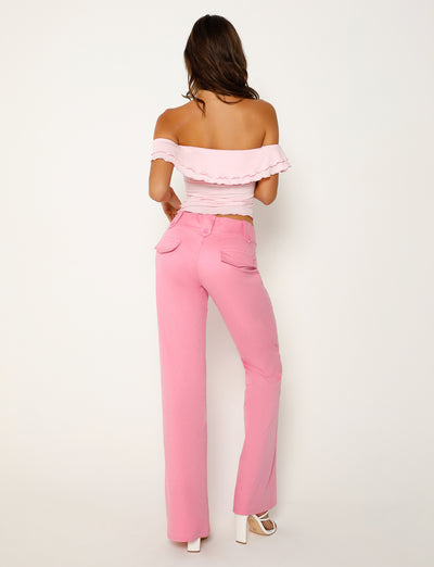 A woman is standing with her back to the camera, facing a white background. She is wearing a pink off-the-shoulder ruffled crop top and high-waisted TIGERMIST KITTIE PANT in pink with pockets. She is also wearing white high-heeled shoes. Her long hair flows down her back.