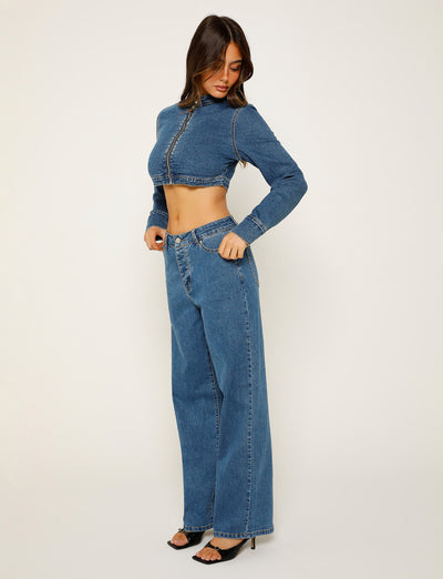 A woman poses against a plain background, wearing the FELIX JEAN - BLUE: DARK WASH from TIGERMIST. She is looking down and to the side, with her left hand resting in her pocket. She wears black, open-toed high heels.