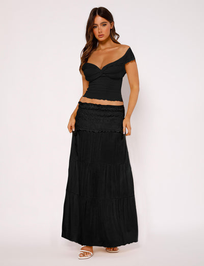 A woman stands against a plain background, wearing the DENALI TOP - BLACK from TIGERMIST along with a long, tiered black skirt. She has long, loose hair and is looking directly at the camera. She is also wearing white open-toe heels.