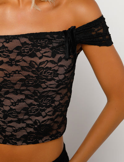 A person wearing the CYD TOP - BLACK from TIGERMIST. This off-the-shoulder, sheer lace top features elegant floral patterns and a small black bow on the left side. The person's right arm is bent, with their hand resting on their hip against a plain white background.