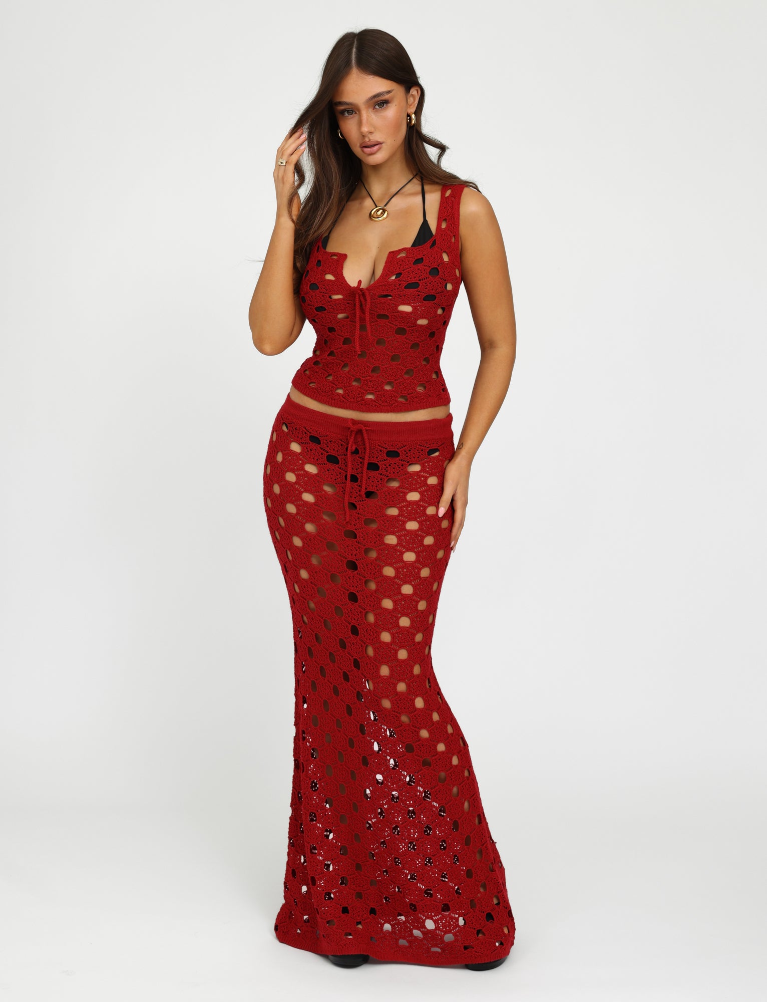 A woman is posing against a plain white background, wearing the Cherry Red Kincade Skirt from Tiger Mist. The top, a red crochet matching piece, is sleeveless and features a deep neckline. The long skirt flows gracefully to her feet with intricate patterns. She has long hair and minimal accessories for a chic look.