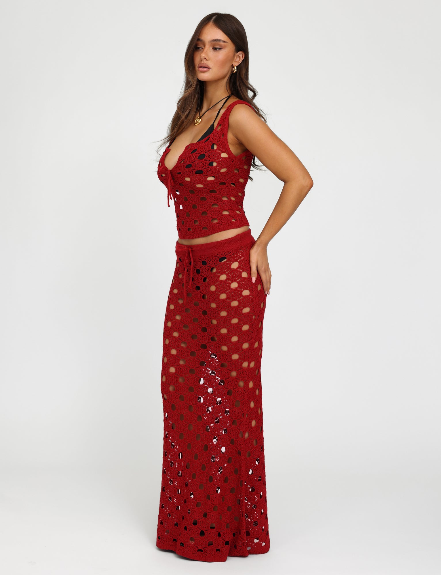 A woman with long, wavy brown hair is wearing the Kincade Skirt in Cherry Red from Tiger Mist. The top is sleeveless with a plunging neckline, and the skirt is long and fitted. She stands against a plain white background, facing slightly to the side with one hand on her hip.