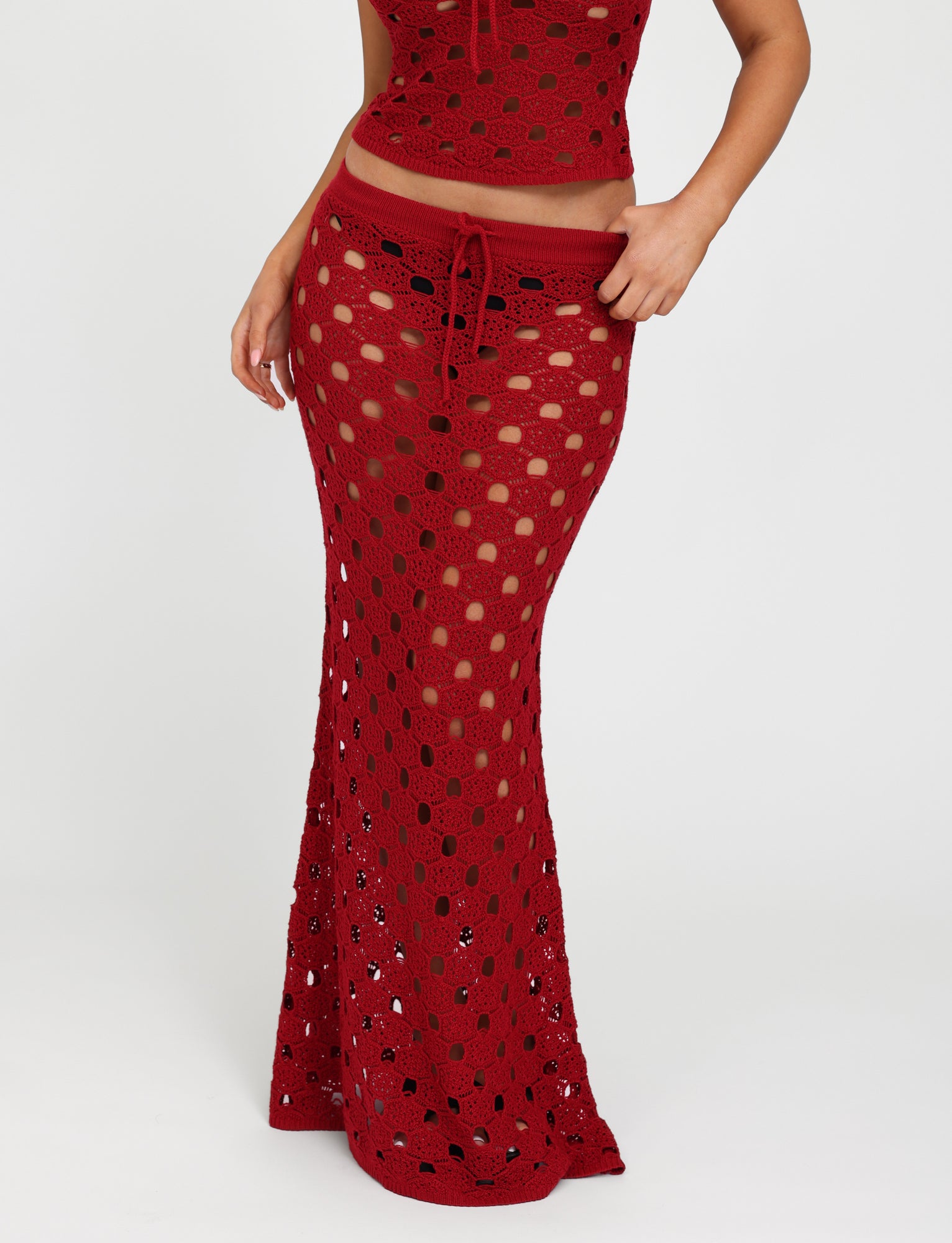 A person modeling the TIGERMIST KINCADE SKIRT in Cherry Red, featuring a red crochet maxi design with a drawstring waist. The fabric has an open, see-through pattern that reveals the legs beneath. The person is also partially showing a matching crochet top, set against a plain white background.