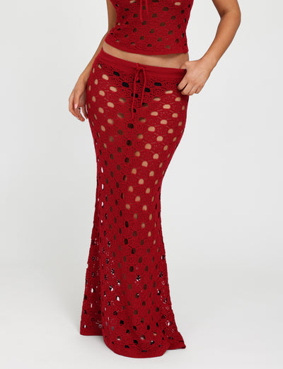 A person modeling the TIGERMIST KINCADE SKIRT in Cherry Red, featuring a red crochet maxi design with a drawstring waist. The fabric has an open, see-through pattern that reveals the legs beneath. The person is also partially showing a matching crochet top, set against a plain white background.