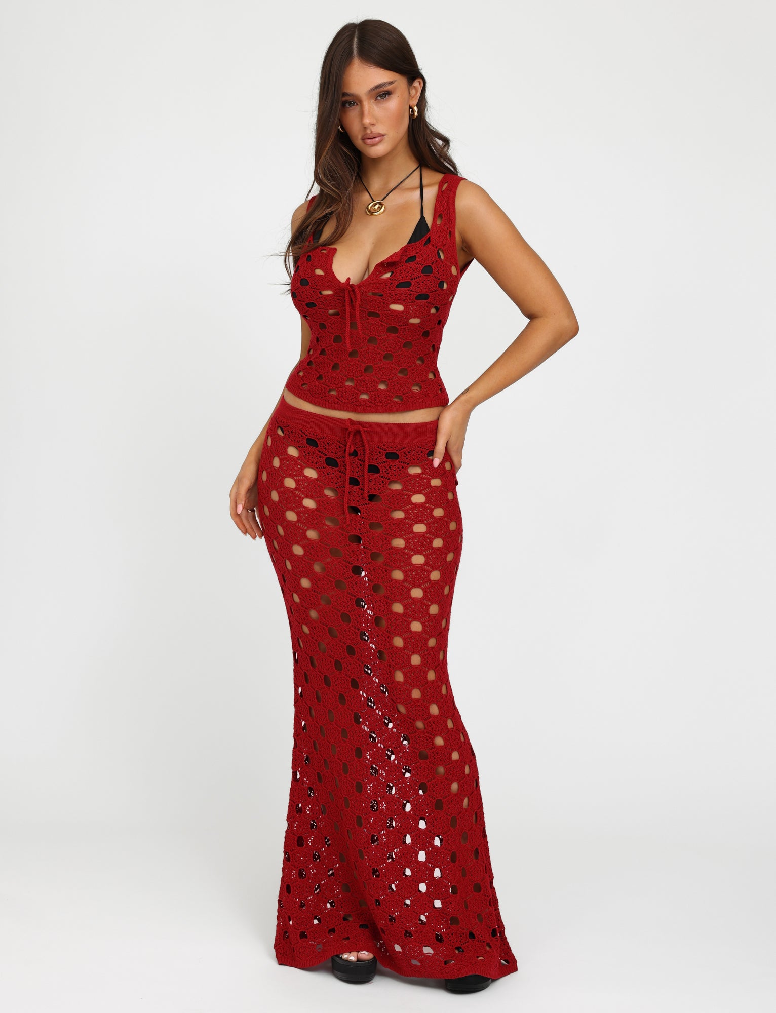 A woman stands against a plain white background wearing the Cherry Red KINCADE TOP from Tiger Mist. The sleeveless top features intricate crochet detailing and circular cutouts. She pairs it with a matching long skirt. Her long, wavy brown hair cascades over her shoulders, and she accessorizes with a necklace and earrings.