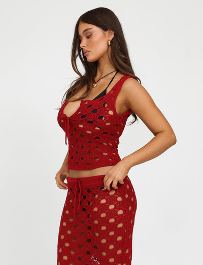 A woman with long dark hair is wearing the TIGERMIST Kincade Top in Cherry Red and a matching skirt featuring circular cutouts. She is facing sideways, looking down, with her left hand resting on her hip against a plain white background.