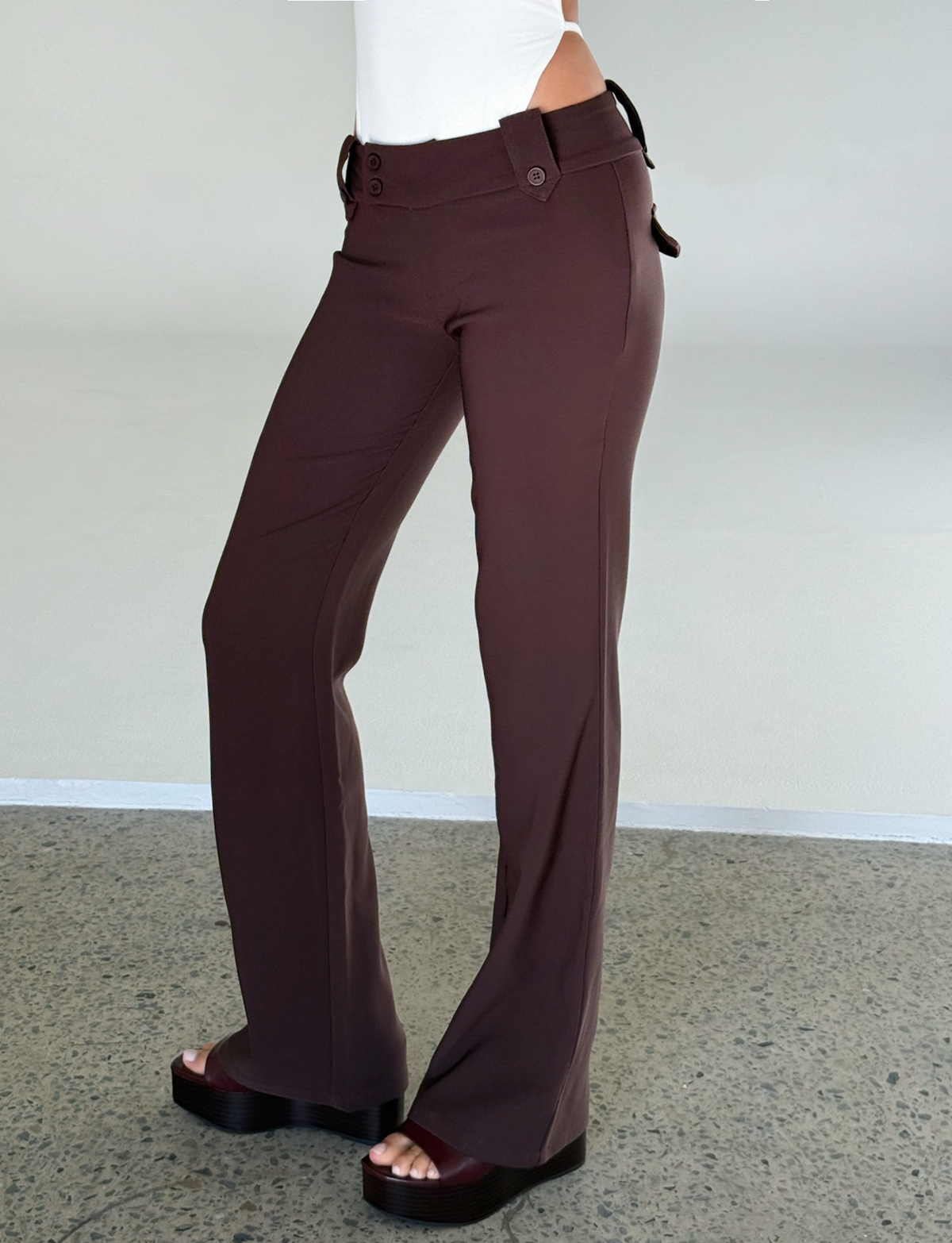 A person in TIGERMIST’s KITTIE PANT - BROWN : CHOCOLATE, featuring a high waist, flared legs, button details, and an asymmetrical waistband, pairs them with black platform sandals and a white top on a textured gray floor.