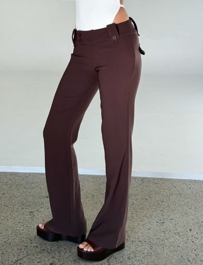 A person in TIGERMIST’s KITTIE PANT - BROWN : CHOCOLATE, featuring a high waist, flared legs, button details, and an asymmetrical waistband, pairs them with black platform sandals and a white top on a textured gray floor.