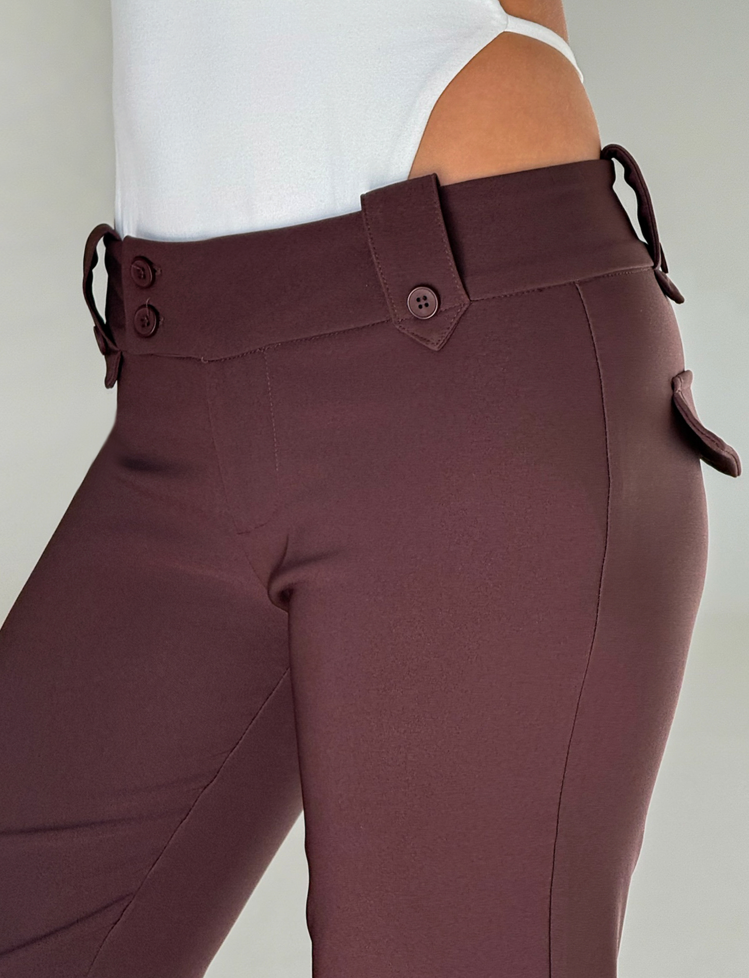 A person wears TIGERMIST's KITTIE PANT - BROWN : CHOCOLATE, a high-waisted, flare fit pant with belt loops and button details, styled with a white top. The image highlights the waist and upper legs.