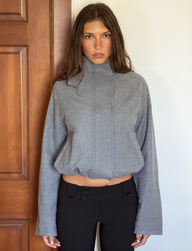 A woman with long brown hair stands indoors, facing the camera in a neutral expression, wearing the TIGERMIST LENNEX JACKET in heather charcoal and black high-waisted pants with a bungee drawstring hem.
