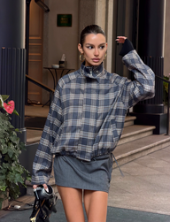 A woman stands outdoors near building steps, wearing the TIGERMIST LENNEX JACKET - GREY CHECK with a short gray skirt, accessorized with a handbag, slicked-back hair, and gold hoop earrings.