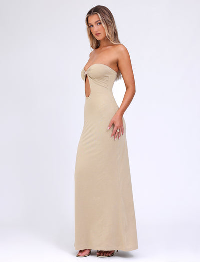 A woman with long hair wears the Cecily Maxi Dress in a strapless style from Tigermist, showcasing a gold colorway with a front cutout and knot detail, paired with brown heeled sandals. She stands in front of a plain white background, facing slightly to the left.