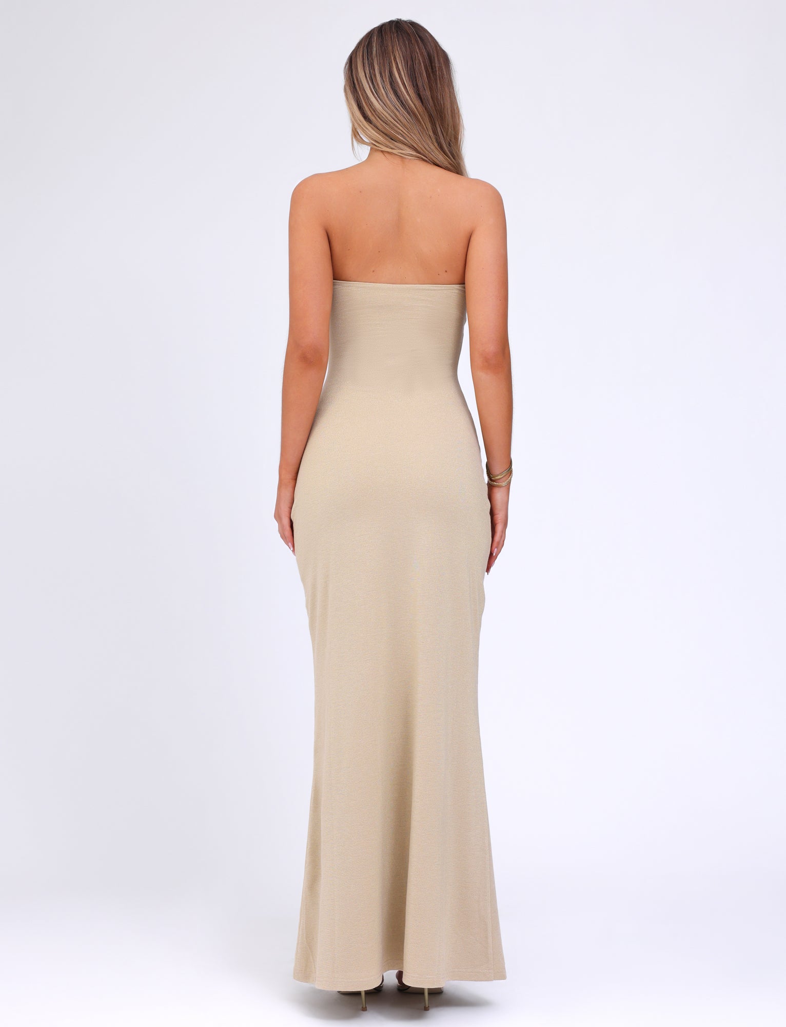 A woman with long hair stands with her back to the camera, wearing a strapless Cecily Maxi Dress in a form-fitting gold gown by TIGERMIST that extends to the floor. She has her left hand resting on her side and her right arm hanging naturally. The background is plain white.