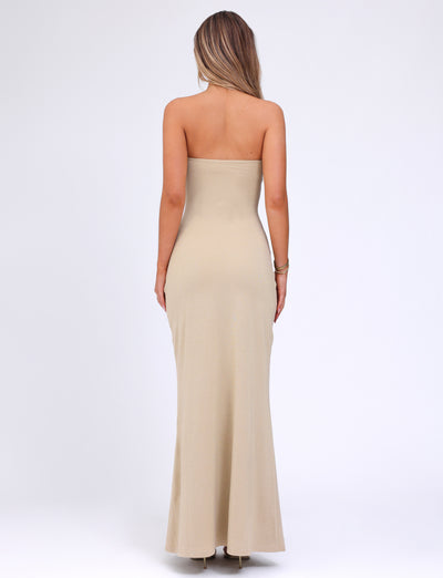 A woman with long hair stands with her back to the camera, wearing a strapless Cecily Maxi Dress in a form-fitting gold gown by TIGERMIST that extends to the floor. She has her left hand resting on her side and her right arm hanging naturally. The background is plain white.