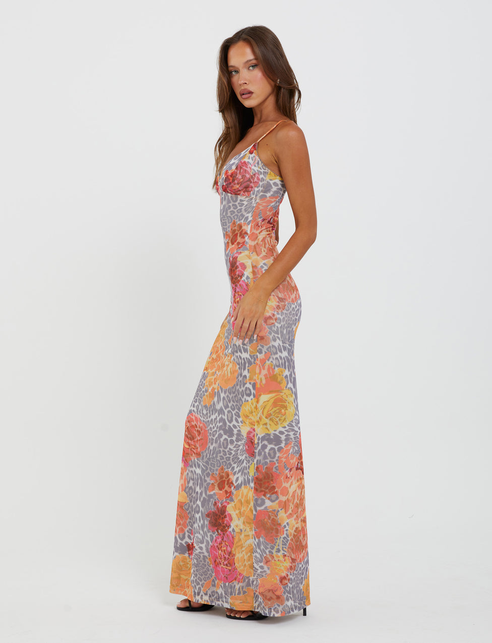A woman stands against a plain white background, wearing the JULIANNA DRESS - MULTI: LEOPARD: ORANGE LEOPARD from TIGERMIST. The long, sleeveless dress with spaghetti straps showcases a vibrant floral pattern in shades of orange, pink, and yellow, accented with gray leopard print. She has long, loose hair and is wearing high heels.