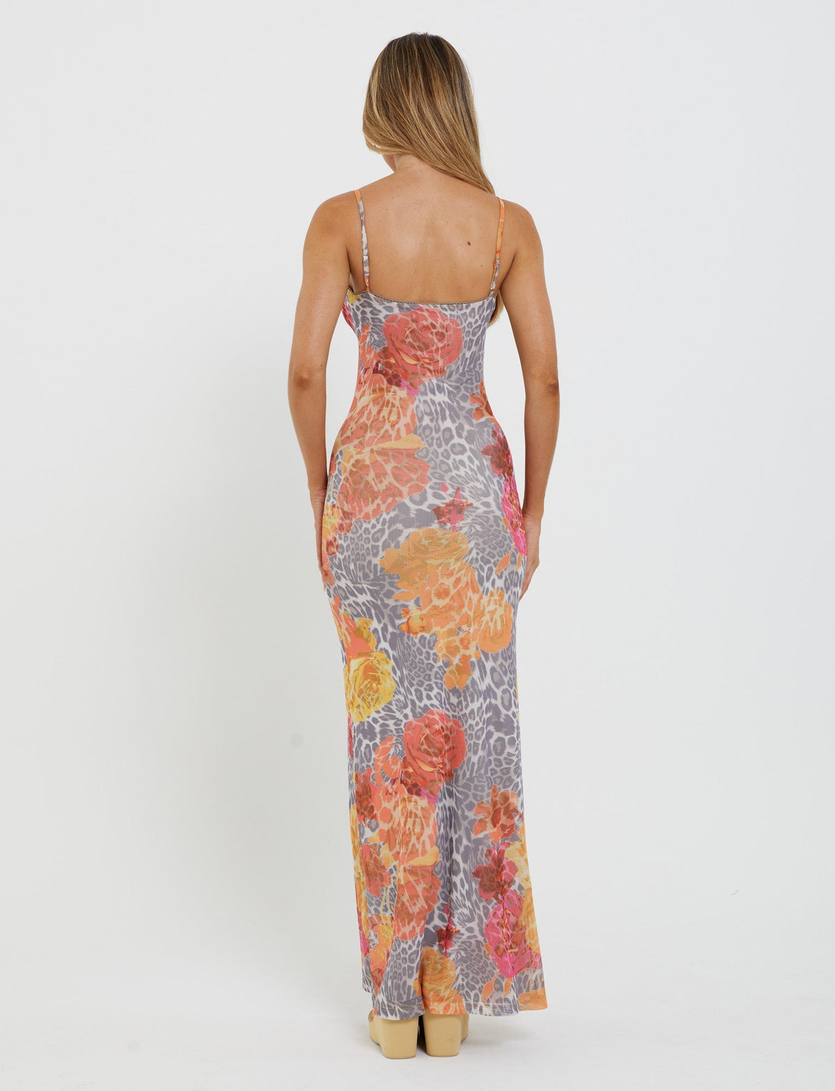 A person with long, light-brown hair is standing with their back to the camera, wearing a sleeveless, form-fitting TIGERMIST JULIANNA DRESS in a multi-colored floral and orange leopard print pattern. The dress is full-length and paired with beige high heels.