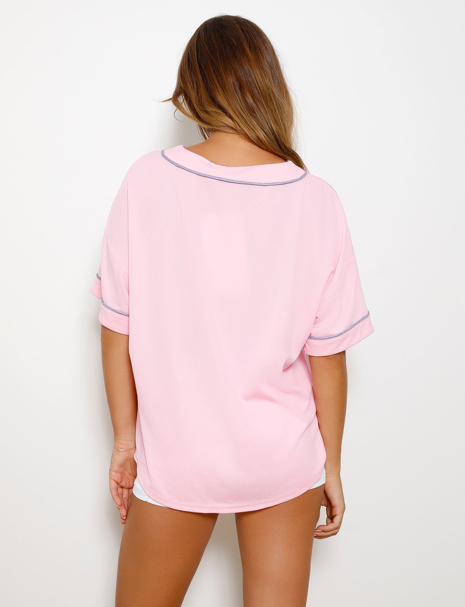 A person with long hair is standing against a plain white background, wearing the TIGERMIST ZEA TOP in Baby Pink. They are facing away from the camera, showcasing the back of this loose-fitting, pastel pink short-sleeved shirt with light gray trim.