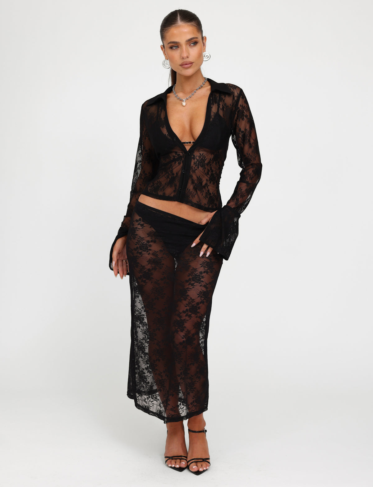 A person stands against a white background wearing the MIRABEL TOP - BLACK : LACE from TIGERMIST, a sheer lace long-sleeve top with a deep neckline, paired with a mid-length skirt. They complete the look with black strappy high heels, hoop earrings, and a necklace. Their hair is styled in a sleek updo.