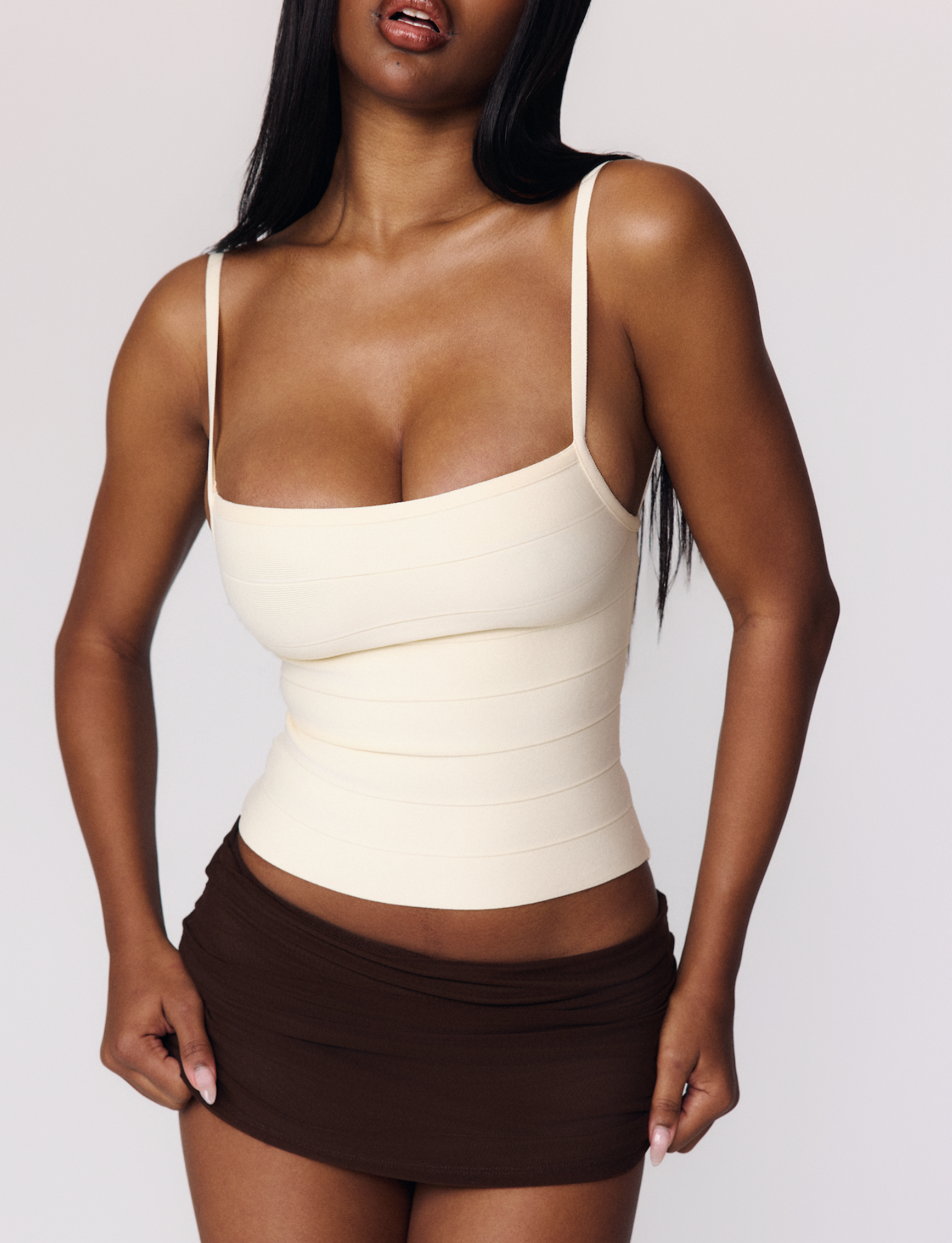 A woman with long dark hair models the TIGERMIST MIDORI BANDAGE TOP in cream, paired with a short brown skirt against a plain light background. Her face is partially out of frame, and her hands rest near the hem of her skirt.