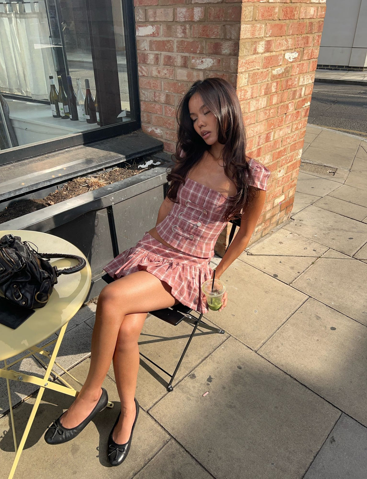 Wearing the TIGERMIST MIKA SKORT in pink plaid, a woman sits at an outdoor table, eyes closed in relaxation as she holds a drink. Sunlight creates patterned shadows on the pavement, and a black bag rests on the yellow table beside her.