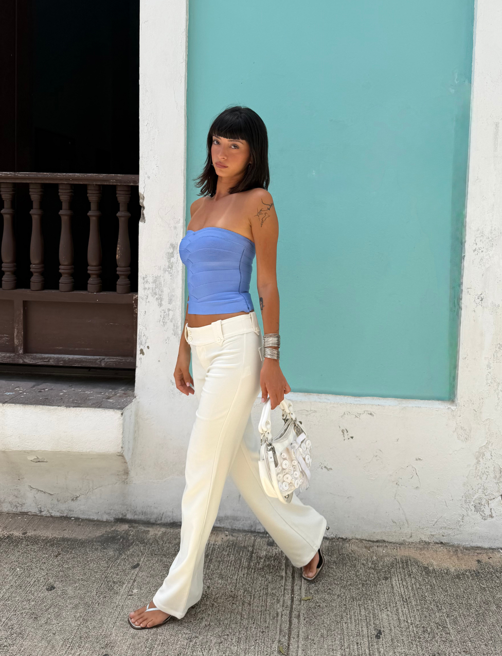 Wearing the TIGERMIST NIYA BANDAGE TOP in Cornflower Blue, a woman with dark hair pairs it with white pants as she walks past a turquoise wall, accessorizing with silver jewelry and carrying a white bag.