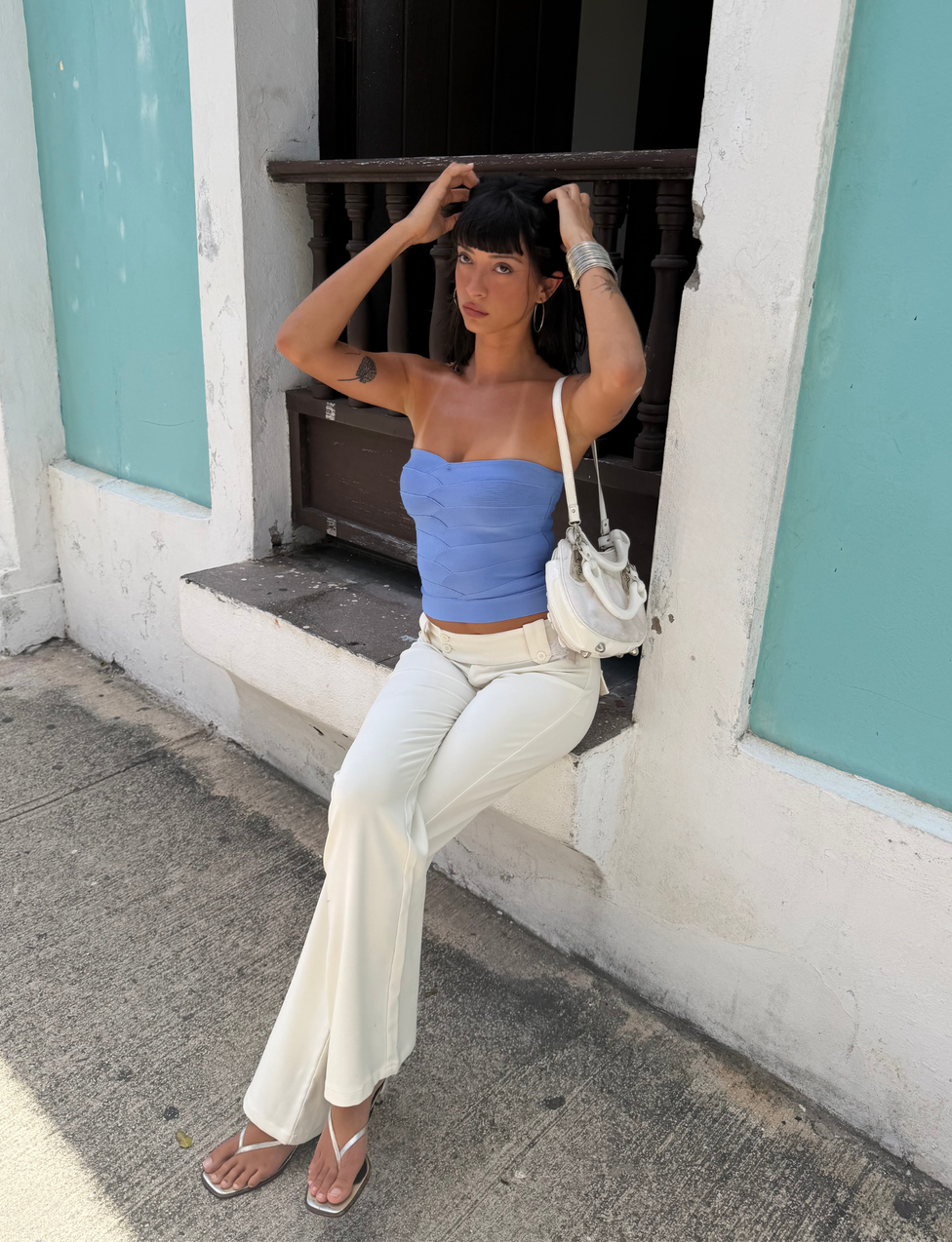 A woman with dark hair, wearing the TIGERMIST NIYA BANDAGE TOP in cornflower blue and white pants, sits on the windowsill of a white and teal building, holding her hair with both hands and carrying a white handbag on her shoulder.